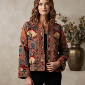May include: A rust-coloured quilted jacket featuring intricate multi-coloured floral embroidery in shades of red, cream, and teal. The jacket has a collarless neckline, long sleeves, and a straight hem, worn over a simple black top and black trousers.