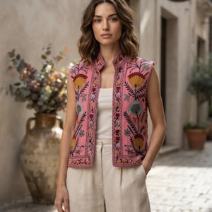 Op de afbeelding: Een mouwloos roze vest met ingewikkeld bloemenborduursel in tinten geel, blauw en grijs. Het vest heeft een hoge kraag, een open voorzijde en wordt gedragen over een eenvoudig wit hemdje in combinatie met een lichtbeige broek met hoge taille.