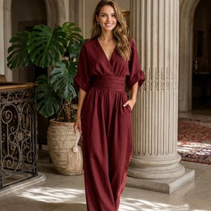 May include: A deep burgundy jumpsuit featuring a V-neckline, elbow-length sleeves with folded cuffs, and a wide, gathered waistband. The garment has long, wide-leg trousers with side pockets and is crafted from a solid-coloured, textured fabric.