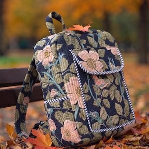 Peut inclure: Un sac à dos matelassé présentant un fond bleu marine foncé avec un motif répétitif de fleurs rose clair et de feuilles vertes. Le sac comprend une poche avant avec un rabat, agrémentée d'un passepoil rayé noir et blanc sur les bords.