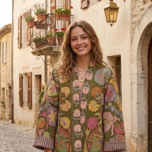 Può includere: Una giacca trapuntata verde oliva con un motivo floreale ricamato in tonalità rosa, giallo e viola. La giacca presenta un'apertura frontale, maniche lunghe con polsini a righe e due tasche anteriori, indossata sopra un top di colore chiaro.