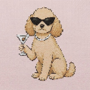 May include: A cross-stitch embroidery on light pink fabric featuring a tan dog wearing black sunglasses and a white beaded necklace. The dog is sitting upright and holding a martini glass with an olive in its right paw, rendered in detailed stitches.