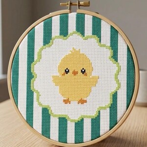 May include: A circular wooden embroidery hoop containing a cross-stitch design of a yellow chick centred on a white scalloped background. The background fabric features vertical teal and white stripes. The hoop has a metal tightening screw at the top.