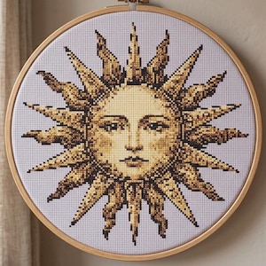 Peut inclure: Un tambour à broder circulaire en bois contenant un motif au point de croix représentant un soleil avec un visage humain. Le soleil est réalisé en fils dorés, marron et noirs sur un tissu blanc, avec des rayons pointus rayonnants.