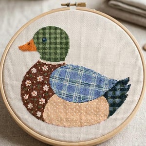 May include: A circular wooden embroidery hoop containing a cross-stitch design of a duck. The duck features a green plaid head, an orange beak, a brown chest with white floral patterns, a blue plaid wing, and a dark green plaid tail.