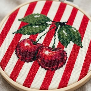 May include: A circular wooden embroidery hoop containing a piece of fabric with a cross-stitch pattern. The fabric features vertical red and white stripes with a central design of two dark red cherries attached to green stems with two green leaves.