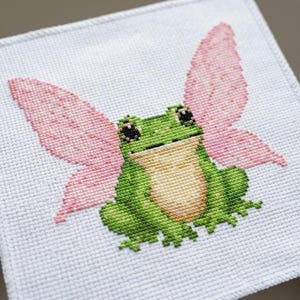 May include: A square piece of white Aida fabric featuring a cross-stitch embroidery of a green frog with a light tan belly and large black eyes. The frog is depicted with two large, textured pink wings extending from its back, creating a fairy-like appearance.