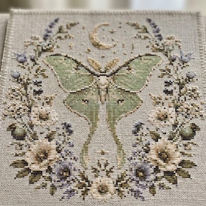 Pode incluir: Um bordado em ponto cruz sobre tecido bege claro apresentando uma mariposa-lua verde-pálida central rodeada por uma coroa circular de flores brancas e roxas, folhas verdes e pequenos botões, encimada por uma lua crescente e estrelas.