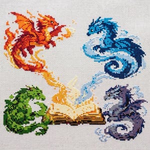 Könnte beinhalten: Eine Kreuzstichstickerei auf weißem Stoff mit vier Elementardrachen, die ein offenes Buch umgeben. Ein orangefarbener Feuerdrache, ein blauer Wasserdrache, ein grüner Erddrache und ein violetter Luftdrache sind mit wirbelnden Energiespuren dargestellt.