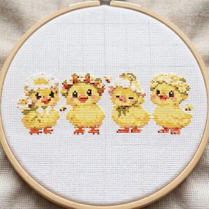 May include: A circular wooden embroidery hoop containing white Aida fabric with a cross-stitch pattern of four yellow chicks. The chicks feature orange beaks and feet, with various floral and eggshell accessories on their heads, arranged in a row.