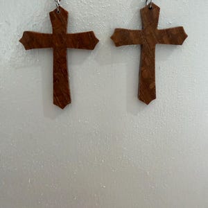 May include: A pair of wooden cross earrings. Each earring features a cross shape with pointed ends. The crosses are a medium brown color and hang from silver-colored fish hook ear wires. The earrings are displayed against a white background.
