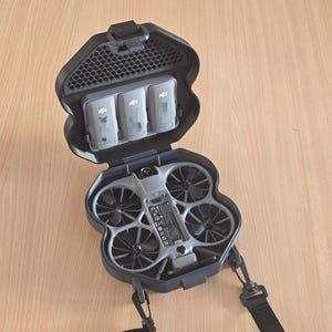 Op de afbeelding: Een zwarte draagtas met een grijze drone met vier propellers en een centrale camera. Het open deksel van de tas bevat drie grijze DJI-batterijen die horizontaal zijn geplaatst. De tas is voorzien van een zwarte verstelbare schouderriem aan de zijkanten.