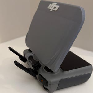 May include: A gray drone remote control with a screen cover. The remote has two black antennas and a power button. The DJI logo is visible on the screen cover.