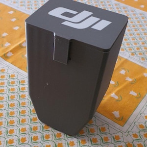 Può includere: Una custodia per batteria DJI grigio scuro di forma rettangolare con un coperchio piatto che presenta il logo DJI bianco. Il contenitore è dotato di un meccanismo di chiusura verticale sul lato anteriore e di una base angolare affusolata in materiale opaco.