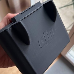 Op de afbeelding: Een matzwart plastic accessoire van het merk DJI met een rechthoekige basis en twee gebogen zijdelingse uitsteeksels. Het oppervlak is voorzien van een gegoten DJI-logo op het voorpaneel. Het object heeft een gladde afwerking.