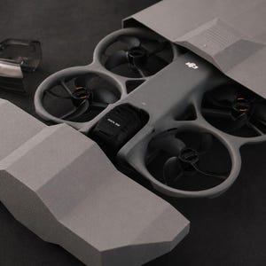 Peut inclure: Un drone gris avec quatre hélices noires est partiellement visible à l'intérieur d'un étui de protection gris. Un couvercle en plastique transparent et une section de l'étui sont séparés. Le drone porte le logo DJI.