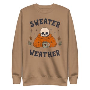 May include: A tan crewneck sweatshirt featuring a graphic of a skull wearing an orange turtleneck sweater and holding a mug with a heart icon. The text "SWEATER WEATHER" is printed in dark blue above and below the illustration with small leaves.