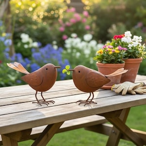 Könnte beinhalten: Zwei rostige Metallvogel-Skulpturen mit ausgebreiteten Flügeln und filigranen Beinen stehen auf einem Holztisch. Einer der Vögel hält einen kleinen grünen Zweig im Schnabel, was auf ein Gartenthema hindeutet. Der Hintergrund zeigt bunte Blumen und Pflanzen.