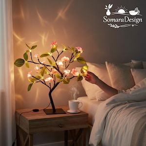 Op de afbeelding: Een decoratieve boomvormige lamp met roze bloesems en groene bladeren. De lamp staat op een houten nachtkastje naast een bed. Een kopje koffie staat op het nachtkastje. De takken van de lamp zijn verlicht.