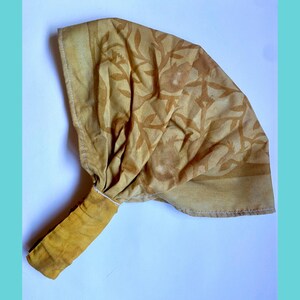 May include: A mustard yellow fabric headwrap featuring a repeating botanical pattern of leaves and pomegranate-like shapes. The accessory includes a gathered section and a wide, flat elasticized band at the base, finished with visible white stitching.