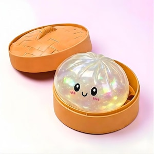 Può includere: Una luce notturna a forma di raviolo perlato con un volto sorridente, all'interno di un vaporiera di bambù arancione. La vaporiera ha un coperchio e lo sfondo è rosa chiaro. La luce notturna emette una luce colorata.