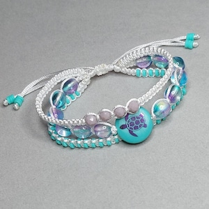 May include: A layered bracelet featuring iridescent beads, turquoise accents, and a central turquoise bead with a purple turtle design. The bracelet is made with white macrame cord and adjustable closures.