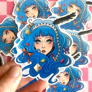 May include: A die-cut sticker featuring an illustration of a person with long, wavy blue hair, green eyes, and a white lace headband. The hair is adorned with a black feather, a small gray mouse, a yellow button, a red lipstick, and a white envelope.