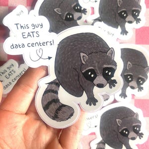 May include: A die-cut vinyl sticker featuring a gray and black raccoon with a striped tail. The sticker includes a speech bubble with the text "This guy EATS data centers!" and a small heart icon with an arrow pointing toward the raccoon.