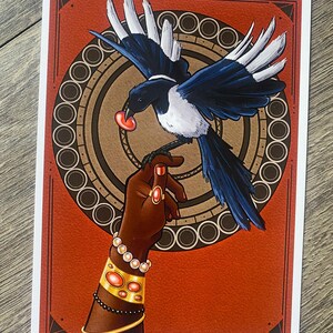 May include: Illustration of a magpie bird with black and white feathers, holding a red object in its beak, perched on a hand adorned with jewelry. The artwork features a red background with a circular design and a decorative border.