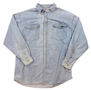 Puede incluir: Una camisa de mezclilla azul claro de manga larga con cuello clásico, cierre frontal de botones y dos bolsillos en el pecho con solapas abotonadas. La camisa presenta mangas largas con puños abotonados y un dobladillo curvo con lavado.