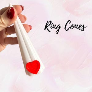 May include: White ring cone with a red heart accent. The cone-shaped jewelry holder has the words "Ring Cones" in black script. The background is a soft pink and white gradient.