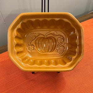 May include: A rectangular ceramic baking dish in a glossy mustard yellow finish. The interior base features a molded relief design of a central pumpkin surrounded by detailed leaves and vines, with scalloped edges along the inner walls.