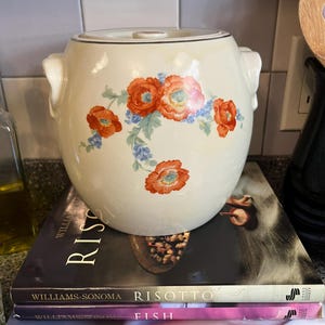 May include: A cream-colored ceramic jar with a lid and two side handles, featuring a floral pattern of orange poppies and small blue flowers. The jar is resting on a stack of four Williams-Sonoma cookbooks titled Risotto, Fish, Wine & Food, and Dinner Parties.