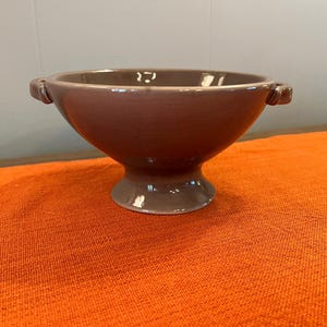 May include: A smooth, glossy, taupe-colored ceramic bowl with a wide, open top and a pedestal base. The bowl features two small, integrated handles on opposite sides, providing a simple, functional design with a uniform, reflective finish.