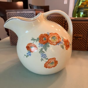 May include: A cream-colored ceramic pitcher with a rounded body and a curved handle. The exterior features a floral pattern of orange poppies and small blue flowers with green leaves. The rim of the pitcher is accented with a thin silver metallic trim.