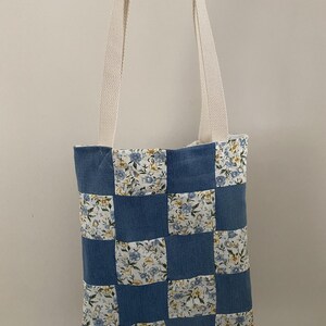 May include: A tote bag featuring a checkered pattern of solid blue fabric squares and floral print fabric squares with light blue and yellow flowers. The bag has two long, off-white woven fabric straps attached to the top edge for carrying.