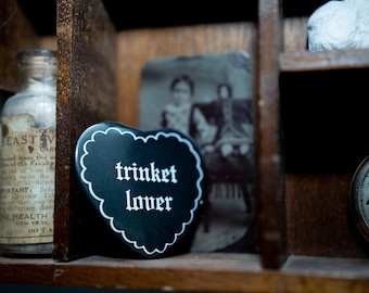 Op de afbeelding: Een zwart hartvormig object met een witte geschulpte rand en de tekst "trinket lover" in witte letters, geplaatst in een houten vak naast een glazen fles met een vintage etiket en een wazige zwart-witfoto.