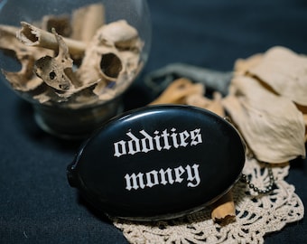 Op de afbeelding: Een zwarte, ovale portemonnee met de witte tekst "oddities money" in een gotisch lettertype. De portemonnee is gemaakt van een glad materiaal en bevat een kleine metalen ketting, bedoeld als functioneel accessoire voor het opbergen van spullen.