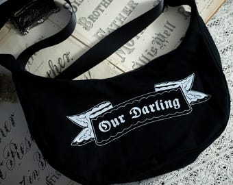 Op de afbeelding: Een zwarte, halvemaanvormige schoudertas met een witte grafische print van een decoratieve banner met de tekst Our Darling. De tas heeft een brede, verstelbare zwarte riem en een gebogen silhouet, gemaakt van een effen zwarte stof.