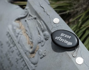Op de afbeelding: Een zwarte, ovale portemonnee met een zilveren kogeltjesketting. De tekst "grave offerings" is in een wit gotisch lettertype op de voorzijde gedrukt. De portemonnee ligt op een grijs stenen oppervlak naast twee kleine zilveren munten.