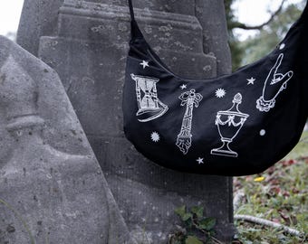 Op de afbeelding: Een zwarte, halvemaanvormige schoudertas met witte lijntekeningen van een zandloper, een decoratieve zuil, een urn met deksel en een handgebaar, geaccentueerd met kleine ster- en zonnesymbolen over het oppervlak van de stof.