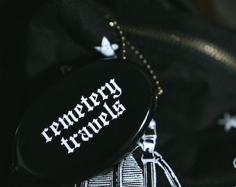 Op de afbeelding: Een zwarte, ovale portemonnee met de witte tekst "cemetery travels" in een gotisch lettertype. De portemonnee is bevestigd aan een zwart stoffen item met een wit sterrenpatroon en een grafische illustratie van een skelet met een camera.