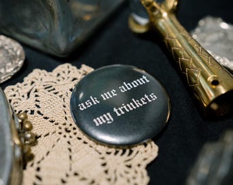 Op de afbeelding: Een ronde zwarte pin-back button met de witte tekst "ask me about my trinkets" in een gotisch lettertype. De button ligt bij een goudkleurig metalen voorwerp met een klauwachtige bovenkant en een beige gehaakt kanten kleedje.