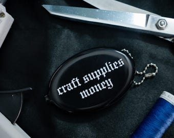 Op de afbeelding: Een zwarte, ovale portemonnee met een zilveren kogeltjesketting, voorzien van de witte tekst "craft supplies money" in een gotisch lettertype. De portemonnee ligt bij een zilveren metalen schaar en een blauwe klos garen op een donkere stof.