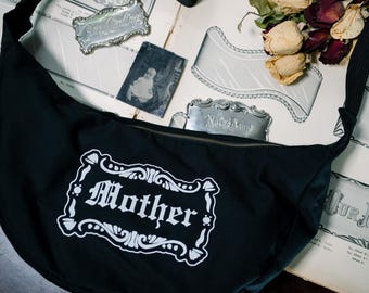 Op de afbeelding: Een zwarte, halvemaanvormige schoudertas met een witte grafische print van een sierlijk rechthoekig kader met het woord Mother in een gotisch lettertype. De tas heeft een lange zwarte schouderband en een ritssluiting aan de bovenkant.