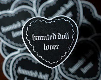 Op de afbeelding: Een zwarte hartvormige sticker met een witte geschulpte rand en de witte tekst "haunted doll lover" in een gotisch lettertype. De sticker ligt over verschillende identieke stickers heen, wat een herhalend patroon van hartjes vormt.