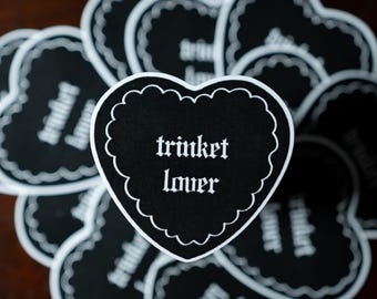 Op de afbeelding: Een verzameling hartvormige stickers met een zwarte achtergrond en een witte geschulpte rand. Elke sticker bevat de tekst "trinket lover" gedrukt in een wit gotisch lettertype, gecentreerd op het oppervlak van de hartvorm.