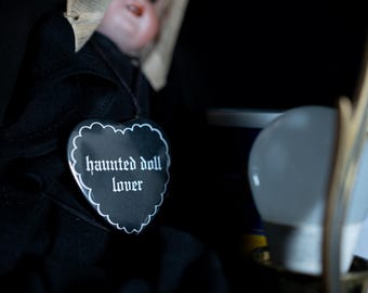 Op de afbeelding: Een pop in zwarte kleding met een beige hoofdbedekking, voorzien van een zwarte hartvormige speld met een witte geschulpte rand en de witte tekst "haunted doll lover" op de voorzijde, geplaatst tegen een donkere achtergrond.