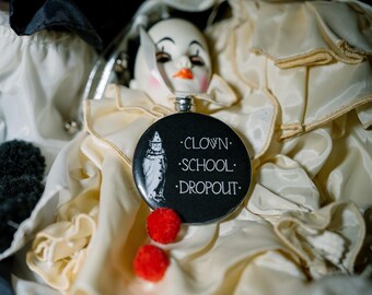 Op de afbeelding: Een ronde zwarte button met een witte illustratie van een clown en de tekst CLOWN SCHOOL DROPOUT in witte letters. De button is versierd met twee kleine rode pompons en is geplaatst op een crèmekleurig satijnen clownskostuum.