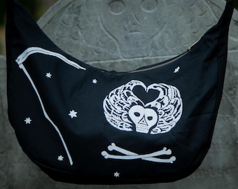 Op de afbeelding: Een zwarte hobo-tas met een witte zeefdruk van een schedel met gevederde vleugels in de vorm van een hart boven gekruiste botten. Het ontwerp bevat een gebogen zeis aan de linkerkant en verschillende kleine sterren verspreid over de stof.